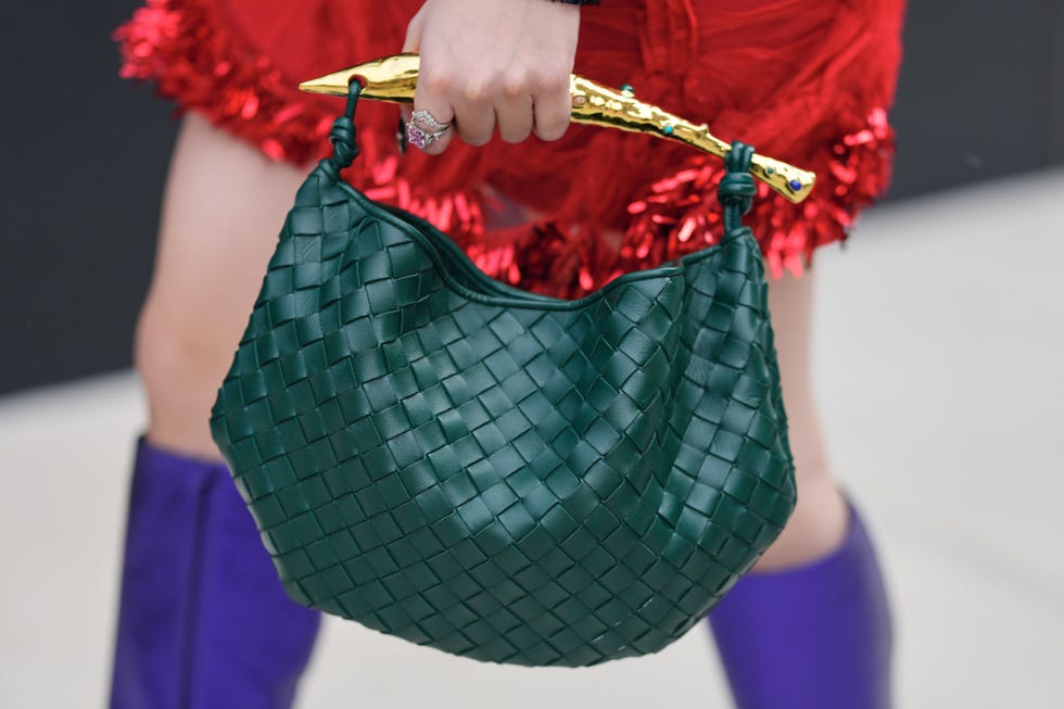 new york, new york september 12: a guest wears white metal rings on the hand, one ring featuring a pink central stone surrounded by clear stones, a slouchy woven dark green leather hobo sardine handbag from bottega veneta with a basketweave pattern, knotted shoulder strap and a sculptural gold tone horn handle set with small blue and green stone studs, a red textured mini skirt trimmed with metallic red fringe, purple knee high boots, outside area, during new york fashion week, on september 12, 2025 in new york, new york (photo by edward berthelot/getty images)
