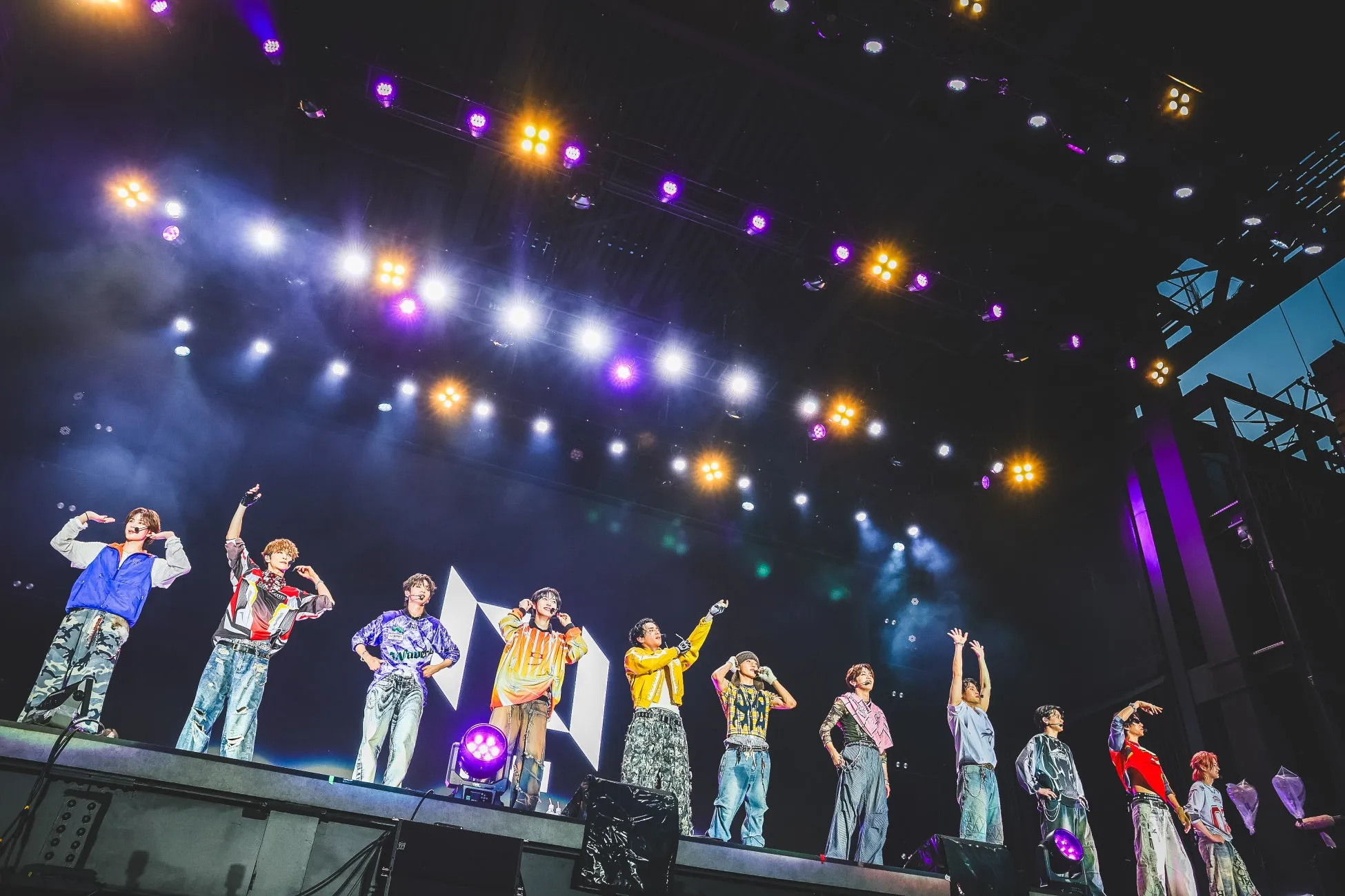 ステージでパフォーマンスをする男性アイドルグループの集合写真。カラフルな衣装を身につけ、照明が輝く中で、メンバーは手を上げたり、マイクを持ったりして観客を魅了している。