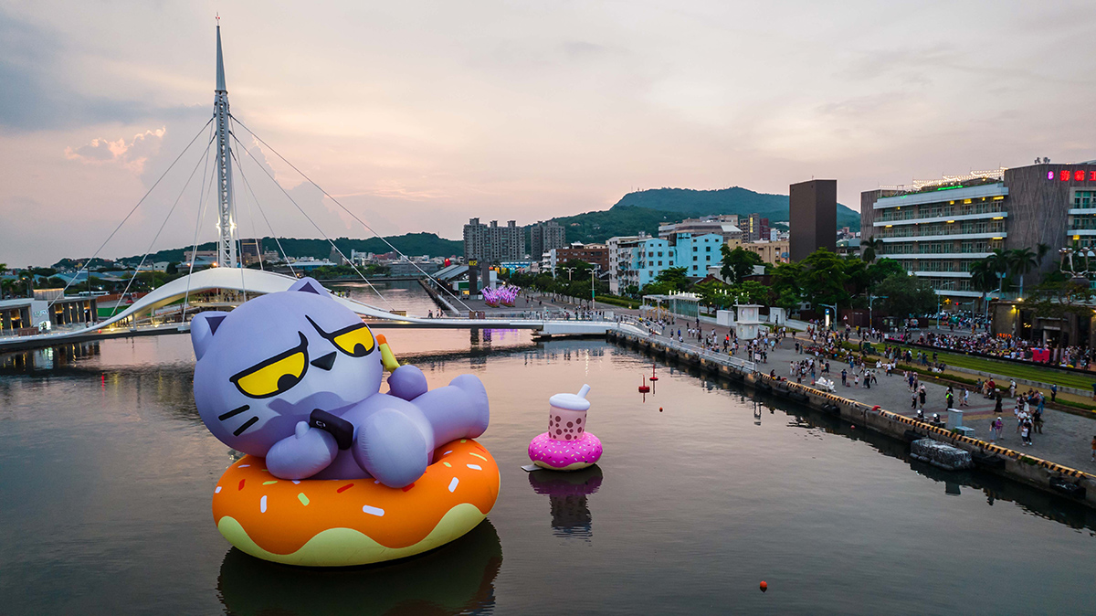 大きな紫のバルーンが水上に浮かび、背景には橋と街並みが広がる夕暮れの風景です。