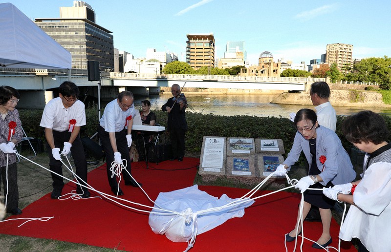 相生橋西詰めの川岸に設置された「原爆の絵碑」の除幕をする関係者。後方は原爆ドーム=広島市で2025年7月22日午後6時12分、佐藤賢二郎撮影