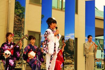 後ろで浴衣を着た4名の演奏でマイクを持って歌っている浴衣姿の女性の写真