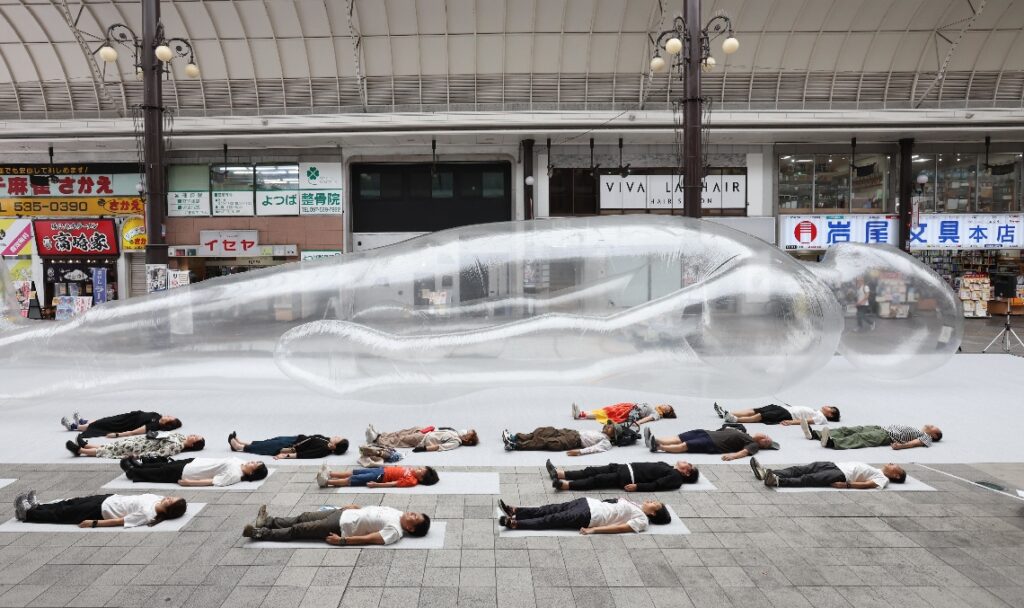 巨大な「空気の人」の横で同じポーズをする人たち＝２７日、大分市のガレリア竹町ドーム広場