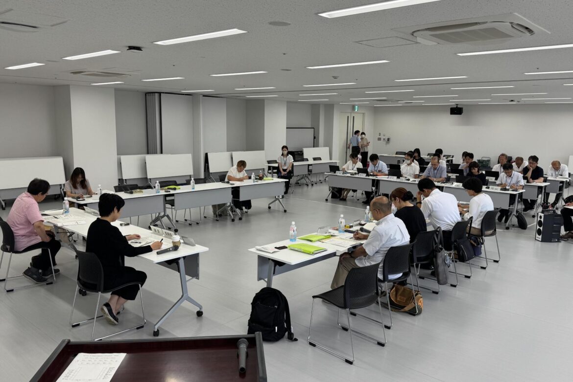 会議室にコの字型に並んだ会議机とその正面に横に数列並んだ会議机の席に出席者たちが座っている委員会の様子の写真