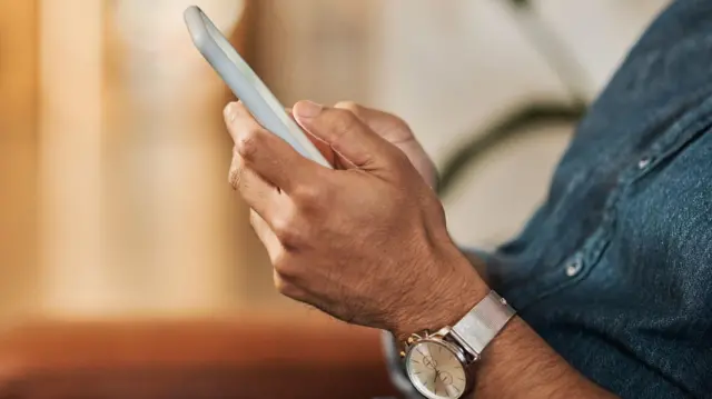 シルバーの腕時計にデニムシャツを着た男性が、両手でスマートフォンを持っている。男性の顔やスマートフォンの画面は見えない