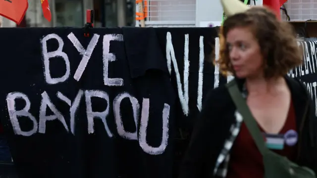 フランス国民議会前の黒いシートに白い塗料で「bye bayrou(バイバイ、バイル) 」と書かれている。その横を女性が通り過ぎている