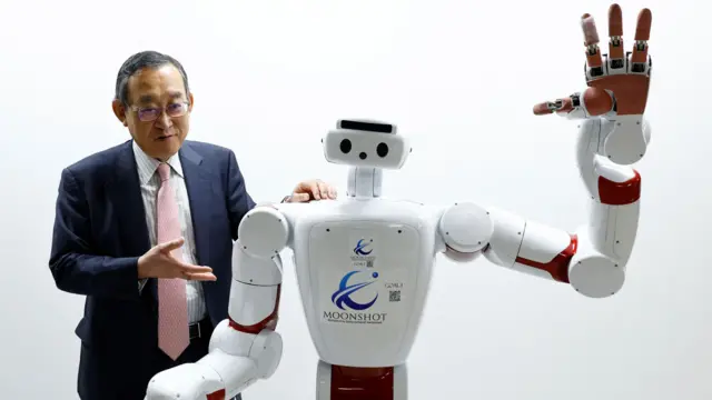 ダークスーツを着てめがねをかけた男性が、白い人型ロボットの隣に立ち、左手をロボットの右肩に乗せている。ロボットは左手を上げている