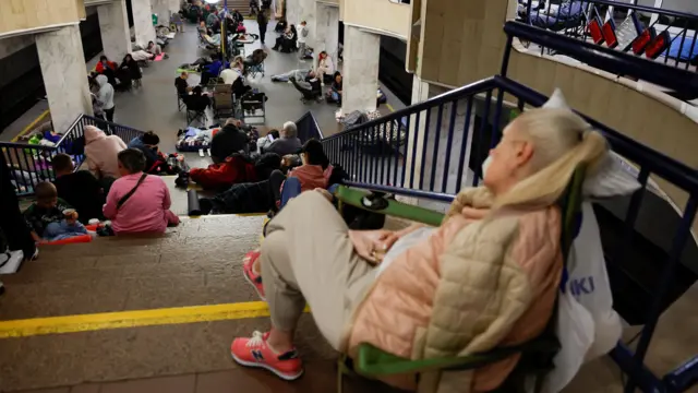 ロシアの攻撃を受け、キーウ市内の地下鉄駅に多くの人が集まっている。1人の女性はベージュとピンクのダウンジャケットを着て、緑の折り畳み椅子にもたれている。構内の階段に腰を掛ける親子や、ホームで横になる人もいる