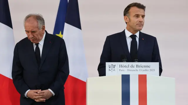 フランス国旗が描かれた演壇で話すマクロン大統領の左隣に、両手を前に組んで足元を見下ろすバイル氏が立っている