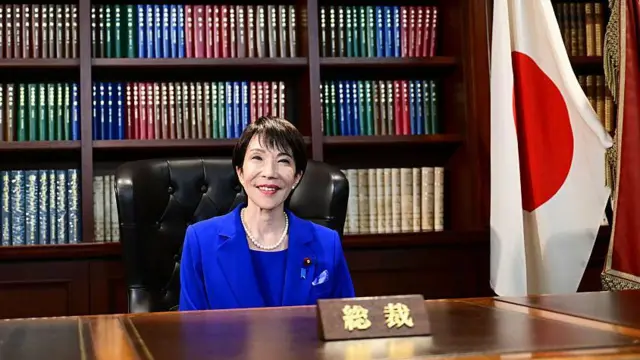 「総裁」と札ののった木の机に向かって座り、笑顔を浮かべる高市氏。本棚と日本国旗などが後ろに並ぶ。総裁選の時と同じ、ロイヤルブルーのスーツを着て、真珠のネックレスとイヤリングをつけている