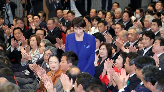 総裁選に勝利し、立ち上がって周りにあいさつする高市氏。周囲は笑顔で拍手している