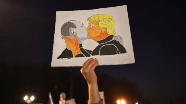 ロシアのウラジーミル・プーチン大統領とアメリカのドナルド・トランプ大統領がキスをしているイラストと、それを掲げる抗議参加者の手と腕の写真