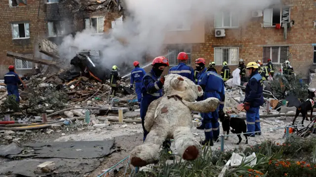 ロシアの攻撃で破壊されたレンガ造りの集合住宅の前にがれきが散乱している。救助隊が黒く汚れた大きな白クマのぬいぐるみを抱えている